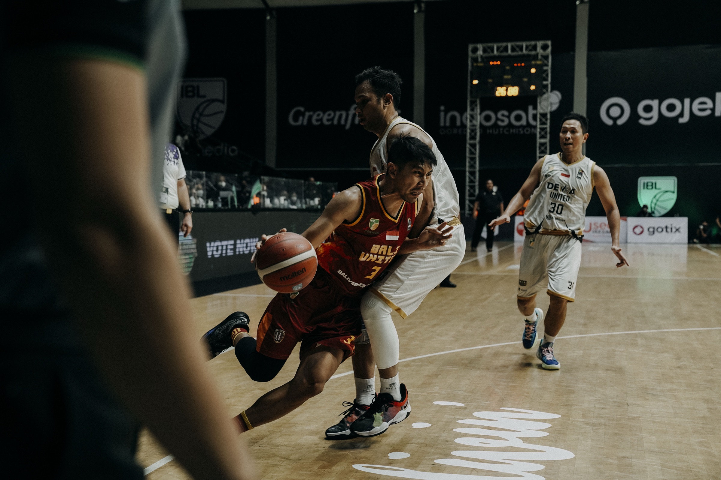 A guard in a dark red Bali United jersey driving past a Dewa United defender during an IBL league game.